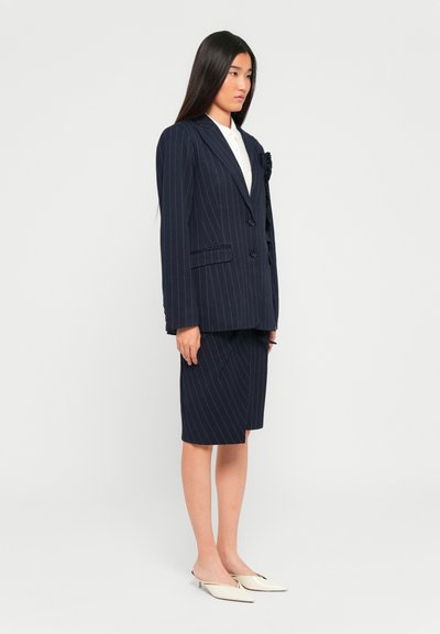 Kvinne iført en marineblå pinstripes blazer og matchende knelangt skjørt med hvite spisse, lave hæler, som står sidelengs mot en hvit bakgrunn.