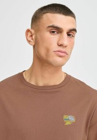 Bruine katoenen T-shirt met een ronde hals, voorzien van een geborduurd logo op de linkerkant van de borst. Korte mouwen en een gladde textuur.