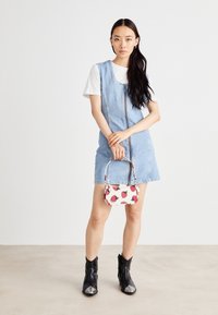 Spijkerjurk met voorrits, korte mouwen en een losse pasvorm. Houdt een kleine witte handtas met rode aardbeien erin vast. Zwarte enkellaarzen.