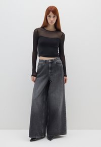Schwarze, durchsichtige Bluse mit langen Ärmeln und gepunkteter Textur, kombiniert mit weit geschnittenen, dunkelgrauen Jeans mit elastischem Bund und unauffälligen Gesäßtaschen.