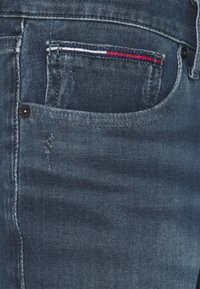 Tmavě modré džínové kalhoty s kapsou, která má červené, bílé a modré prošívání. Zahrnují viditelné opotřebení poblíž okraje kapsy.