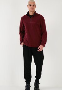 Pullover in fleece di colore bordeaux con colletto a mezzo zip e maniche lunghe, abbinato a joggers neri e stivali neri con lacci. Texture liscia.