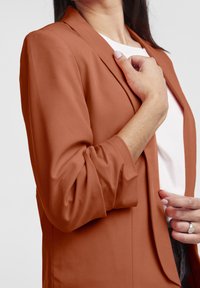 Rostfärgad kavaj med skräddarsydd passform, långa ärmar och en jämn, mjuk textur. Noterbart är den enkla knappstängningen och den klassiska slagdesignen.