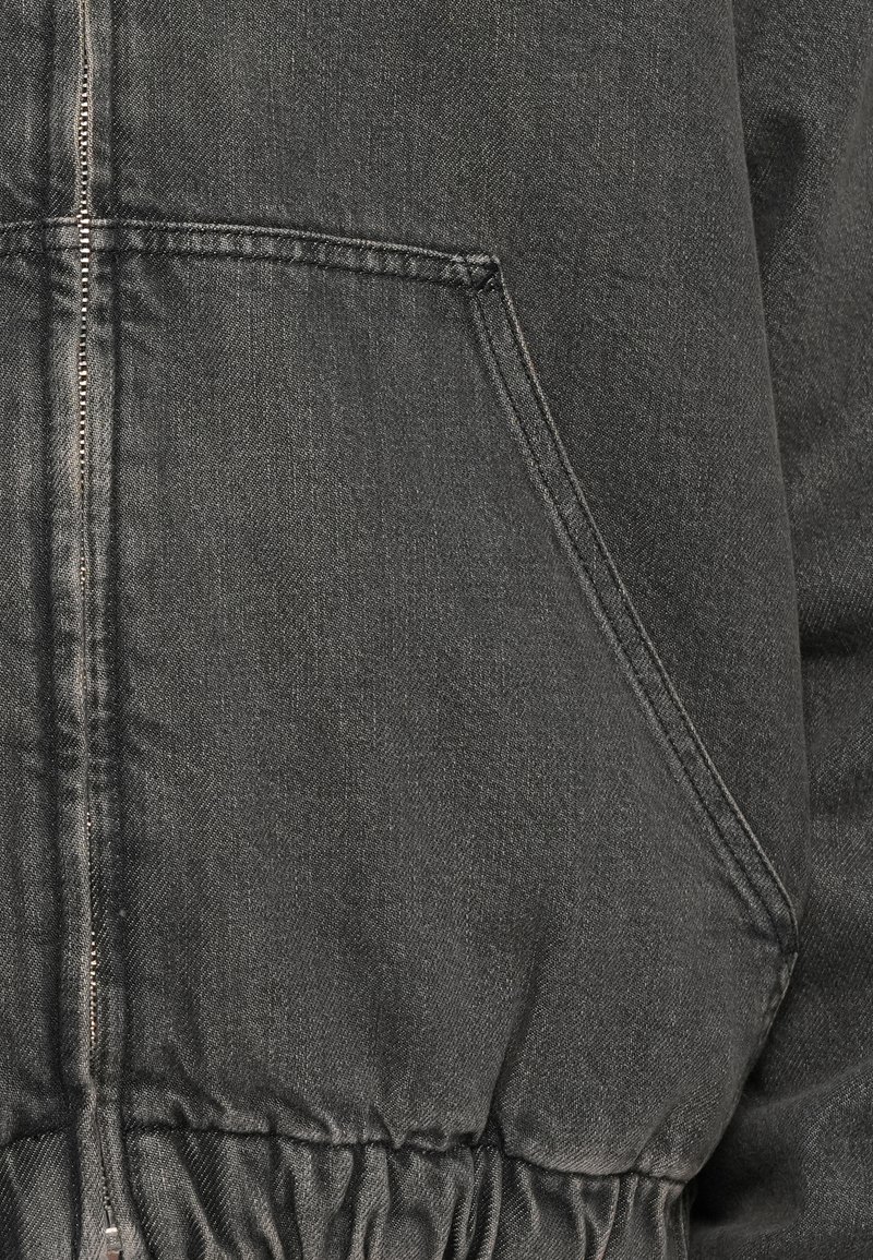 Chaqueta de mezclilla negra con cierre de cremallera, bolsillos laterales y cintura elástica. La tela tiene un acabado texturizado con sutiles detalles de costura.