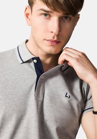 Szara koszulka polo z granatowym kołnierzykiem, z dwoma guzikami z przodu i małym granatowym logo na lewej piersi. Krótkie rękawy z granatowymi akcentami.