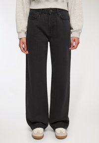 Jeans larghi neri in denim, con vita alta, cinque tasche e un silhouette pulita, abbinati a sneakers chiare.