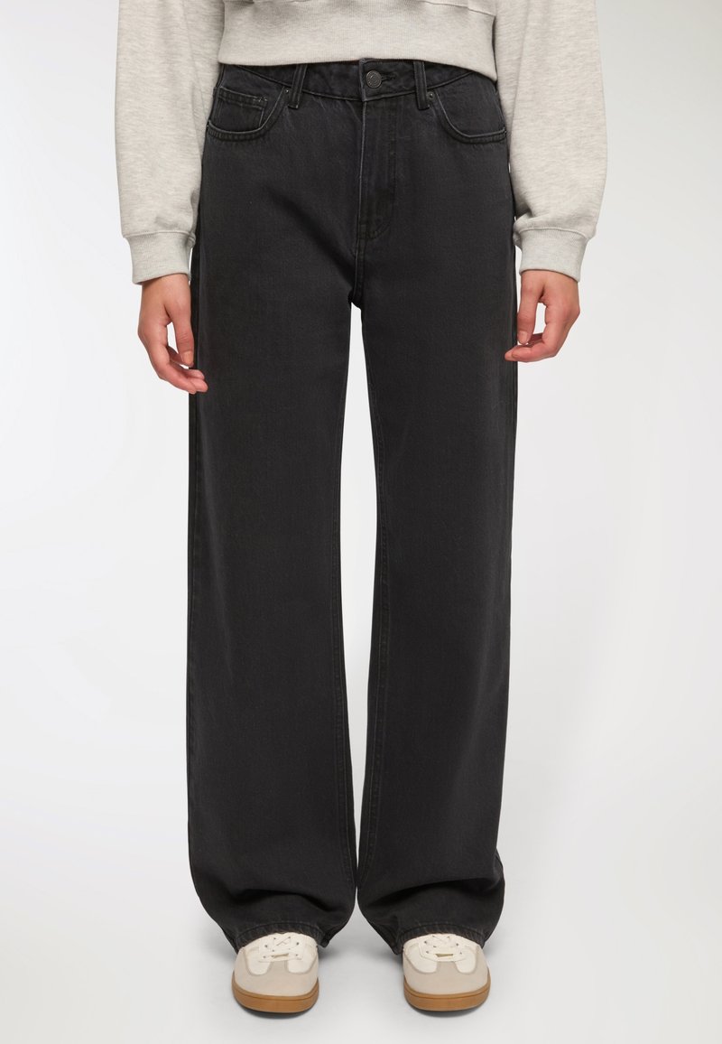 Jeans larghi neri in denim, con vita alta, cinque tasche e un silhouette pulita, abbinati a sneakers chiare.