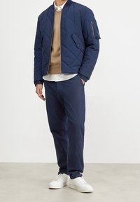 Tmavě modrá prošívaná bomber bunda s žebrovaným límcem, béžový svetřík s kulatým výstřihem a tmavě modré chinos kalhoty. Outfit doplňují bílé tenisky.