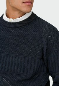 Pull en maille navy avec des motifs texturés en diamant et en côtes. Encolure ras du cou avec un col blanc visible en dessous.