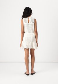 Robe blanche en dentelle avec un col rond, dos ouvert avec un détail en forme de trou de serrure, jupe évasée et motifs texturés. Portée avec des sandales noires.