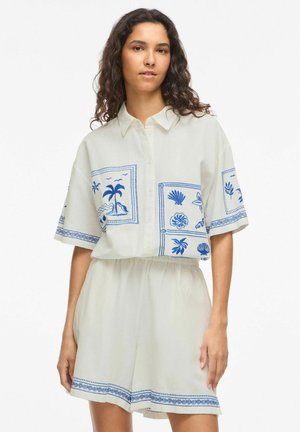 Žena s dlhými kučeravými vlasmi, oblečená v bielej krátkorukávej košeli na gombíky a zodpovedajúcich kraťasoch s modrými tropickými vzormi.