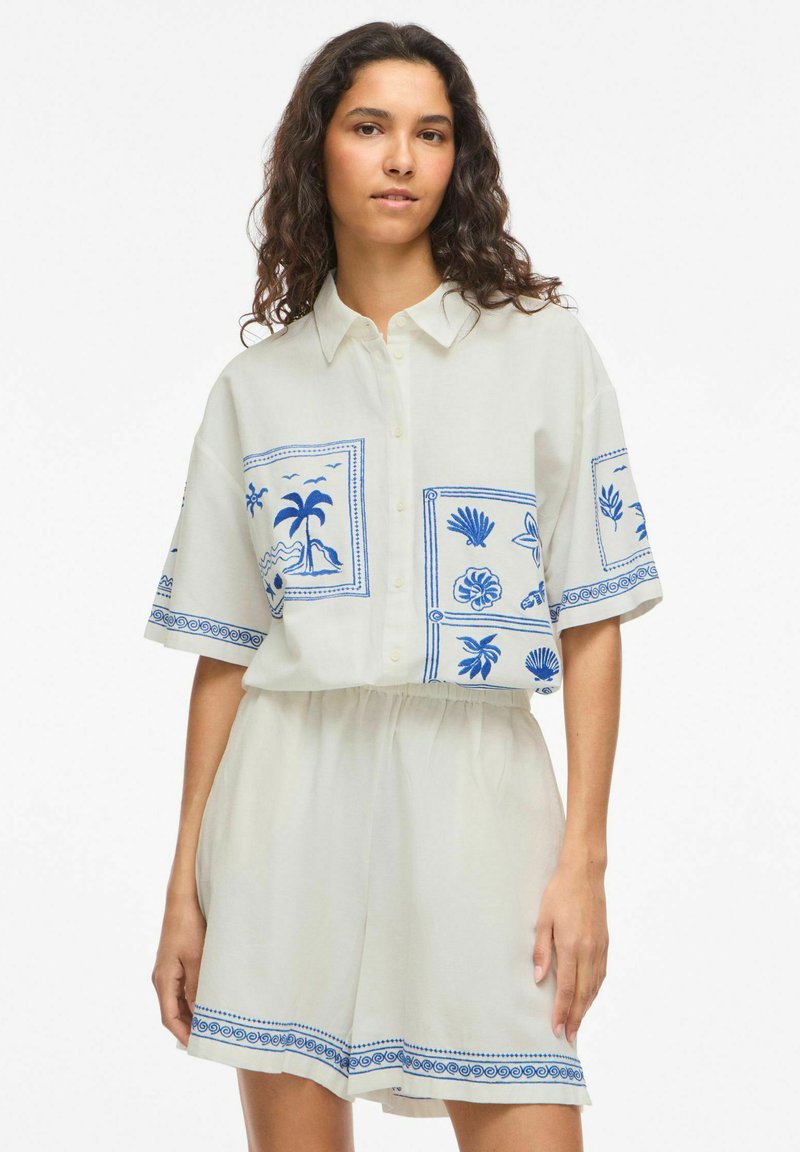Femme aux longs cheveux bouclés portant une chemise blanche à manches courtes boutonnée et un short assorti avec des motifs tropicaux bleus.