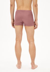 Homme debout pieds nus, de dos, portant un boxer mauve sur fond blanc uniforme.