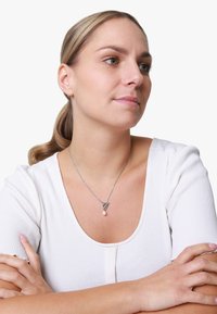 Collana d'argento con un ciondolo a nodo decorativo e una goccia di perla, indossata sopra un top bianco a costine con scollatura rotonda.