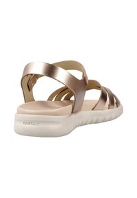 Sándal de bronce metálico con múltiples correas, una suela beige acolchada y una suela blanca texturizada. La marca es visible en el talón.