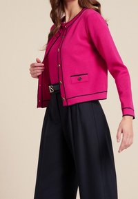 Fuchsiový cardigan s černými detaily, zlatými knoflíky a malými předními kapsami, kombinovaný s širokými námořnickými kalhotami. Hladká textura, strukturovaný design.