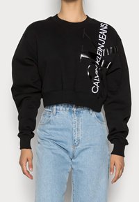 Osoba v černém zkráceném svetru s logem "Calvin Klein Jeans" na rameni a v světle modrých džínech s vysokým pasem.