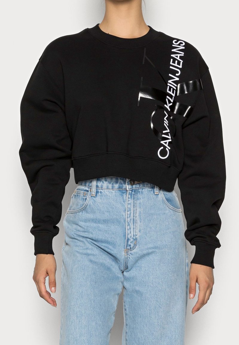 Osoba v černém zkráceném svetru s logem "Calvin Klein Jeans" na rameni a v světle modrých džínech s vysokým pasem.