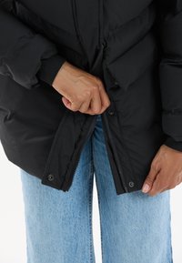 Hænder zipper en sort polstret jakke over lyseblå denimjeans mod en hvid baggrund.