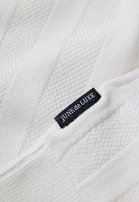 Witte textuurstof met een zwart rechthoekig label erop genaaid, met de tekst "JUNIk de LUXE" in witte letters.