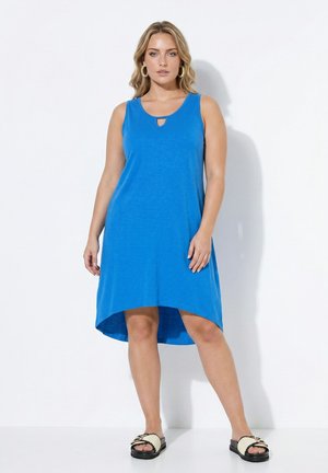 Donna che indossa un vestito blu senza maniche a taglio asimmetrico con scollo a goccia e sandali beige, in piedi davanti a sfondo bianco.