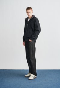 Μαύρο μπουφάν με φερμουάρ και ψηλό γιακά, συνδυασμένο με σκούρα τζιν με ρίγες στο πλάι και λευκά αθλητικά παπούτσια με μαύρες λεπτομέρειες.