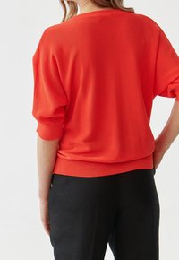 Czerwony dziergany sweter z luźnymi rękawami i prążkowanym dołem, noszony na czarnych spodniach, prezentujący teksturowany, żywy materiał.