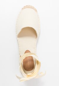 Espadrille en toile blanc cassé avec un bout rond et des rubans à la cheville. Dispose d'une semelle texturée et de coutures le long du bord.