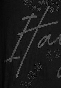 Černé bavlněné tričko s šedým potiskem textu různých písem a velikostí. Design zahrnuje stylizovaná slova a tvary, což vytváří efekt vrstev.