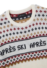Gestrickter Pullover mit cremefarbenem Grund und roten, schwarzen sowie goldenen Rautenmuster. Schwarzer Text mit der Aufschrift "APRÈS SKI" in der Mitte. Rundhalsausschnitt, weiche Textur.