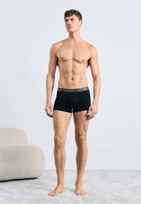 Sorte bomulds boxershorts med en mærket elastik i livet. Modellen har en toneret fysik og står barfodet på en lys struktureret overflade.