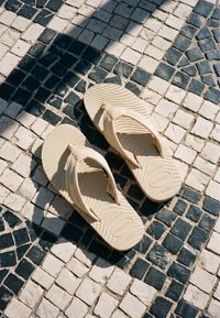 Μπεζ σαγιονάρες Havaianas που ξεκουράζονται σε μαυρο-λευκό πλακόστρωτο με μοτίβο, με αντιθέσεις φωτός και σκιάς.