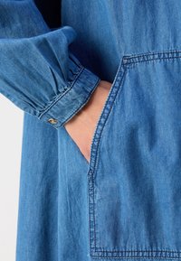 Main insérée dans une grande poche avant d’un vêtement en denim bleu avec un poignet froncé et un détail de bouton sur la manche.