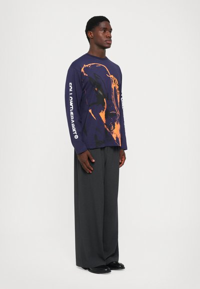 Navy long-sleeve t-shirt featuring an abstract orange graphic; paired with loose, gray trousers and black shoes.