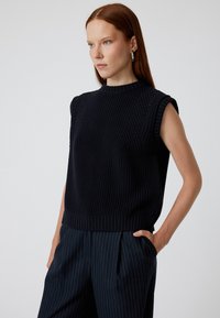 Marinblå stickad ärmlös topp med ribbad textur, rundad hals och lätt sänkta ärmhål. I kombination med rutig byxa.