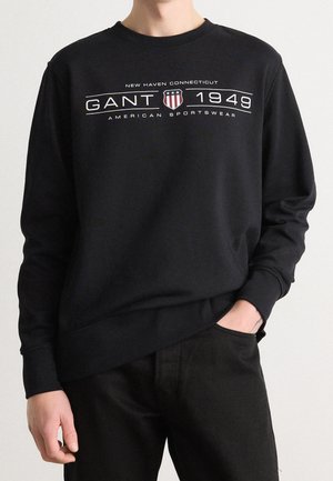 Osoba w czarnej bluzie z kapturem z logo "GANT 1949", herbem tarczy oraz napisem "New Haven Connecticut American Sportswear."