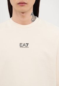 Világos bézs, rövid ujjú póló, puha pamutból készült. A mellkason fekete EA7 Emporio Armani logó található. Sima textúra, klasszikus fazon.