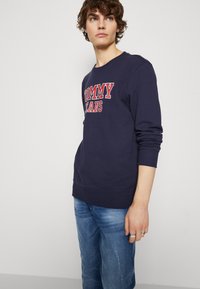 Sudadera marina con el logo rojo "TOMMY JEANS", cuello redondo, puños acanalados y dobladillo inferior. Combinada con jeans desgastados de color azul claro.