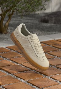 Baskets Tommy Jeans beiges avec semelle en gomme, lacets serrés, exposées au-dessus de carreaux en bois carrés et de petits cailloux gris en extérieur.