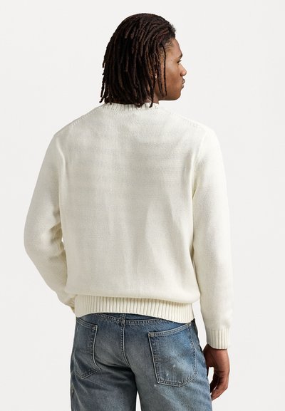 Fehér, kötött pulóver ribbelt ujjal és aljjal, textúrált mintával, kék farmernadrág felett viselve, hátulról nézve.