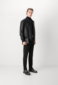 Veste en cuir noir à col, associée à un pull col roulé noir, un pantalon noir ajusté et des chaussures noires à lacets. Tient une pochette noire.