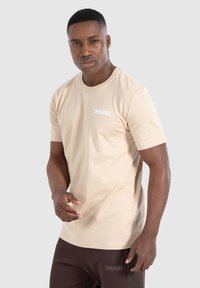 Beige T-shirt met korte mouwen van katoen, met een ronde halslijn en een wit logo op de borst, gecombineerd met donkerbruine broek.