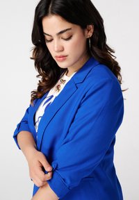Blazer bleu en tissu lisse, avec une coupe ajustée, des manches retroussées avec un détail de bouton, associé à un haut blanc et un collier.
