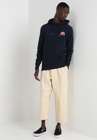 Sudadera azul marino con bolsillo frontal y logo, combinada con pantalones recortados beige claros y zapatos negros sin cordones, de pie contra una pared.