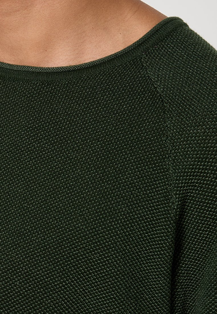 Donkergroen gebreide trui met een brede halslijn, voorzien van een gemêleerd patroon en raglanmouwen. De textuur lijkt enigszins ruw.