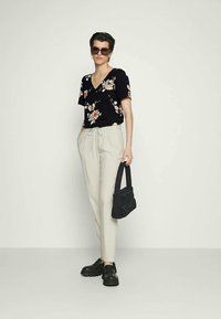 Mulher a usar uma blusa preta com padrões florais, calças bege com cordão, sapatos chunky pretos, óculos de sol, segurando uma mala preta contra um fundo claro.