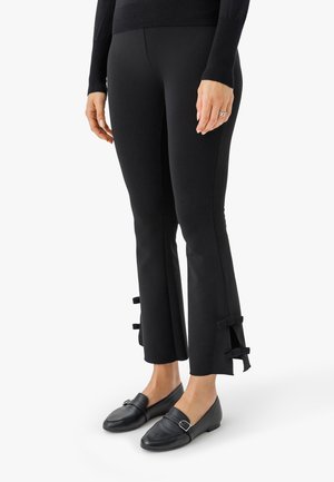 Leggings negras con puños acampanados que presentan pequeños lazos en los lados. Combinados con mocasines de cuero negros con una hebilla decorativa.