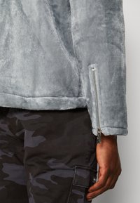 Szara pluszowa kurtka o gładkiej fakturze, z bocznym zamkiem i detalami na mankietach. Połączona z czarnymi spodniami w kamuflaż z kieszeniami typu cargo.