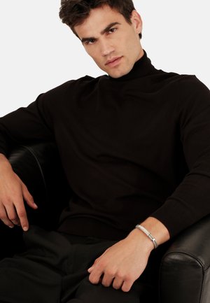 Zwarte coltrui met lange mouwen, gestructureerde stof en een getailleerd ontwerp, gecombineerd met donkere broek en een zilveren armband.
