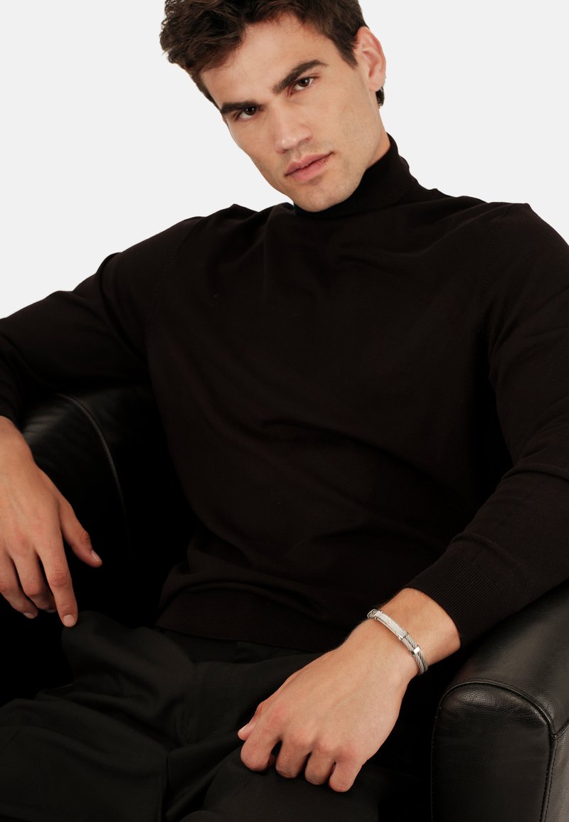 Jersey negro de cuello alto con mangas largas, tejido texturizado y diseño ajustado, combinado con pantalones oscuros y una pulsera de plata.